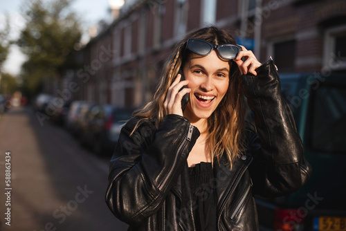 Attractive happy woman talking on cell phone, raised her glasses in surprise, open mouth. Young cute tourist blonde woman with two thin braids outside, wearing black leather jacket and black dress.