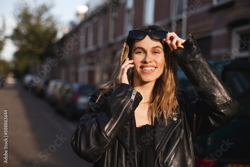 Attractive happy woman talking on cell phone, raised her glasses in surprise, open mouth. Young cute tourist blonde woman with two thin braids outside, wearing black leather jacket and black dress.