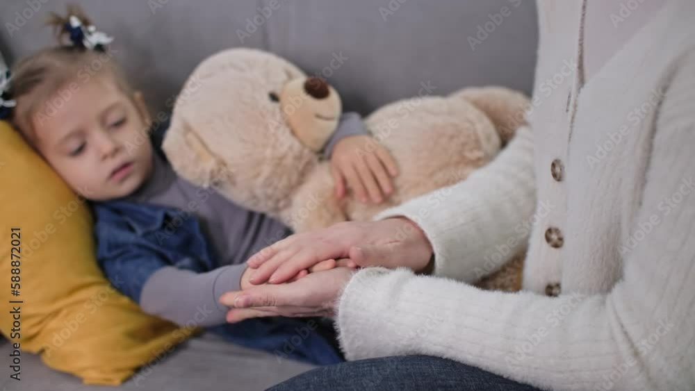 support for parents of children, caring woman stroking the hand of her unhealthy little girl lying on sofa in room support for parents of children, caring woman stroking the hand of her unhealthy little girl lying on sofa in room