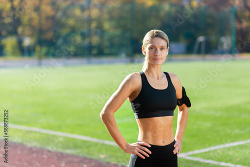 Portrait of mature adult female athlete in stadium, female athlete holding hands on hips smiling and looking at camera, jogging woman in tracksuit during active exercise and fitness class.