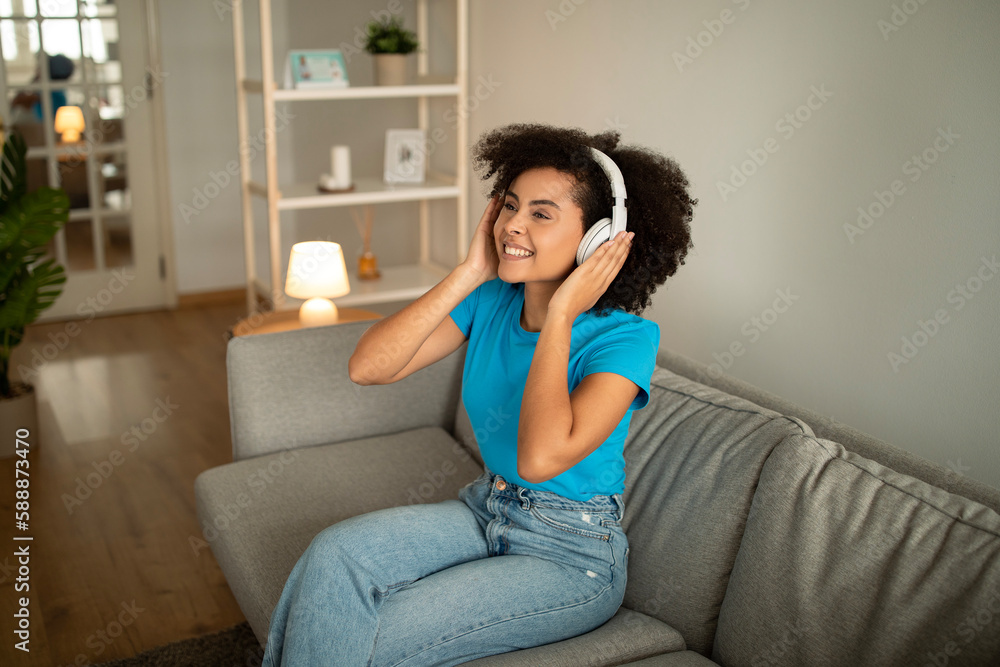 Happy millennial african american curly lady in wireless headphones listen music, enjoy free time on sofa