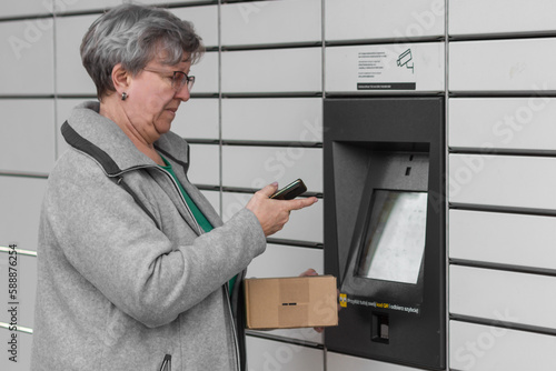 Senior woman with smartphone entering code to open shopping locker in inPost delivery service for receives or sends box.
