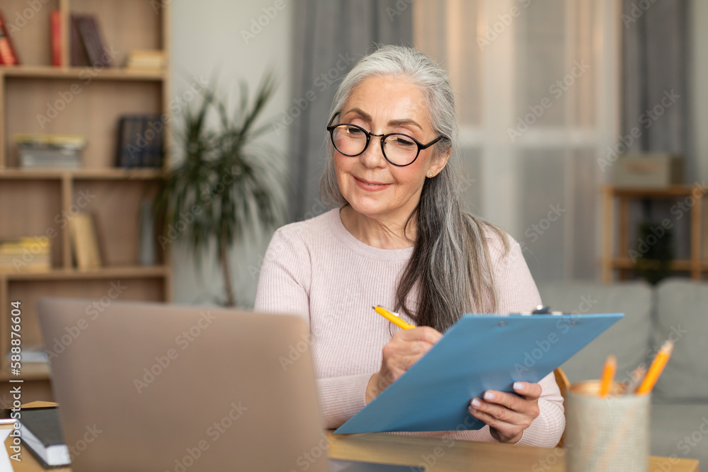 Cheerful caucasian old female in glasses sits at table, makes notes, looks at computer, has video call Cheerful caucasian old female in glasses sits at table, makes notes, looks at computer, has video call