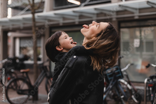 Happy young woman have fun with cute child boy holding him on arms and hug. Mommy little kid son together outdoors. Mother's Day love family. Woman with boy go to home. Mom and son show tongue, play. © zvkate