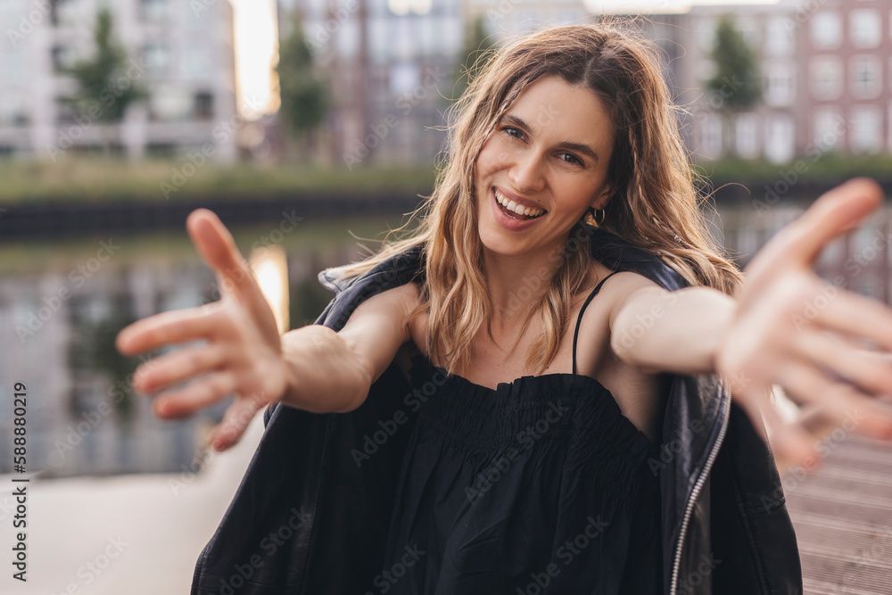 Cheerful lady making come to me sign. Pretty young girl with curly blonde hair and two thin braids look to camera, smiling and wear black leather jacket and dress. Woman invite someone or want hugging