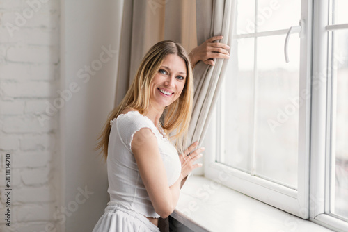 Happy millennial caucasian blonde woman in domestic clothes pulls back curtain, looks out window