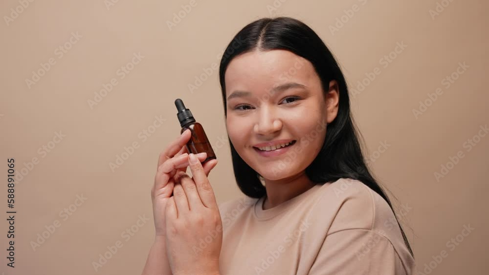 Young brunette woman showing a bottle of serum on a beige background. Beauty skincare commercial. Young asian girl with silky skin face. High quality 4k footage Young brunette woman showing a bottle of serum on a beige background. Beauty skincare commercial. Young asian girl with silky skin face. High quality 4k footage
