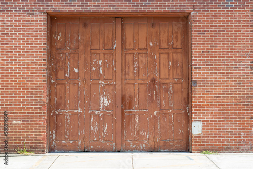 The exterior of a vintage brown brick building with two wooden closed garage doors. There's a pattern of panels on the warehouse doors. The reddish paint is peeling on the double doors exposing grey.