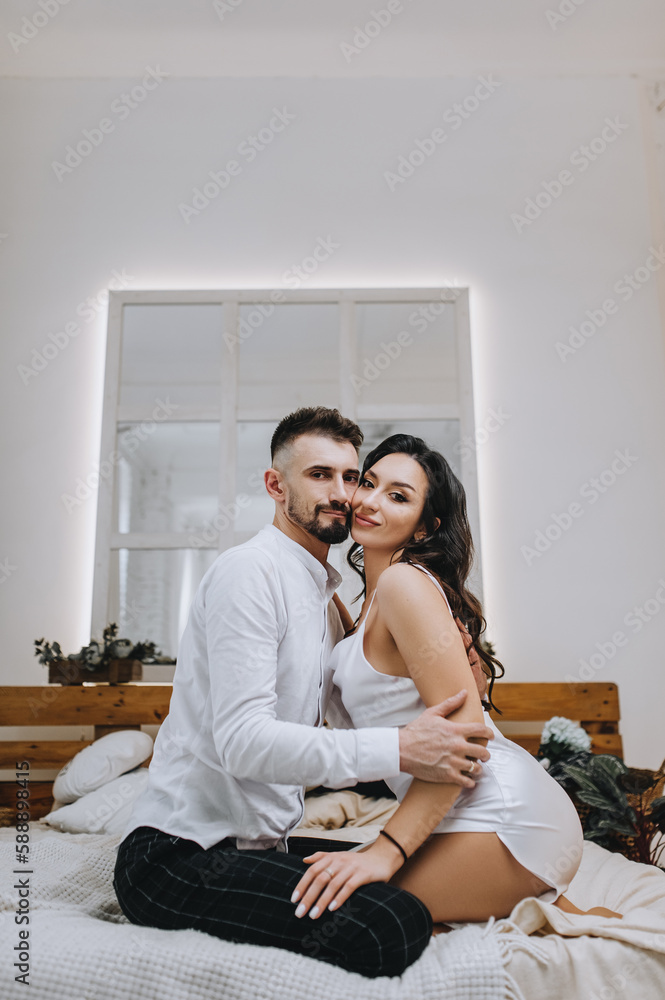 Stylish young bearded groom and beautiful smiling brunette bride with bouquet hugging while sitting on bed in interior, indoors. Wedding photography of happy newlyweds, close-up portrait. Stylish young bearded groom and beautiful smiling brunette bride with bouquet hugging while sitting on bed in interior, indoors. Wedding photography of happy newlyweds, close-up portrait.