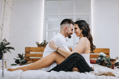 Stylish young bearded groom and beautiful smiling brunette bride with bouquet hugging while sitting on bed in interior, indoors. Wedding photography of happy newlyweds, close-up portrait.