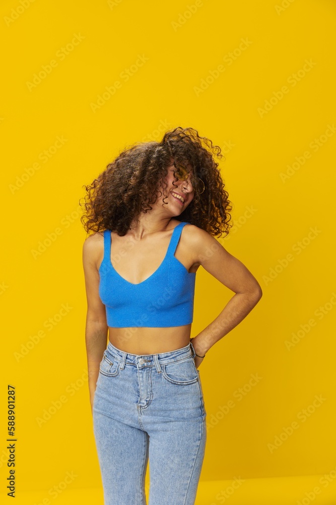 Woman with curly afro hair in a blue T-shirt on a yellow background dancing flying hair with sunglasses yellow, hand signs, look into the camera, smile with teeth and happiness, copy space Woman with curly afro hair in a blue T-shirt on a yellow background dancing flying hair with sunglasses yellow, hand signs, look into the camera, smile with teeth and happiness, copy space