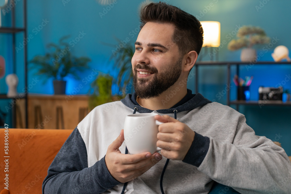 Attractive smiling bearded man drinking a cup of warm coffee or herbal tea in the morning. Portrait of guy enjoying comfortable relaxing taking a break alone at home apartment living room on sofa Attractive smiling bearded man drinking a cup of warm coffee or herbal tea in the morning. Portrait of guy enjoying comfortable relaxing taking a break alone at home apartment living room on sofa
