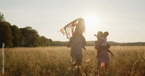 Happy young smiling family of four have fun in a golden wheat field at sunset. Mom, dad, son and daughter walk together holding colorful kite during golden hour.