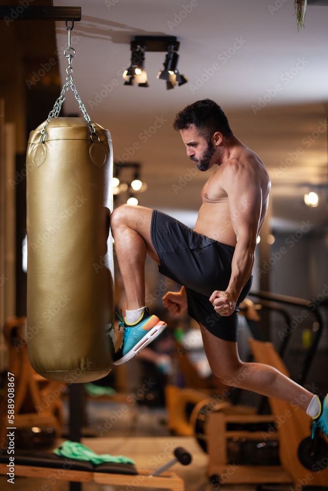 Muscular shirtless man jump hitting a punching bag with a knee in a fitness studio.