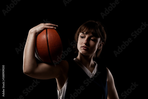 Female basketball player. Beautiful girl holding ball. Side lit half silhouette studio portrait against black background.