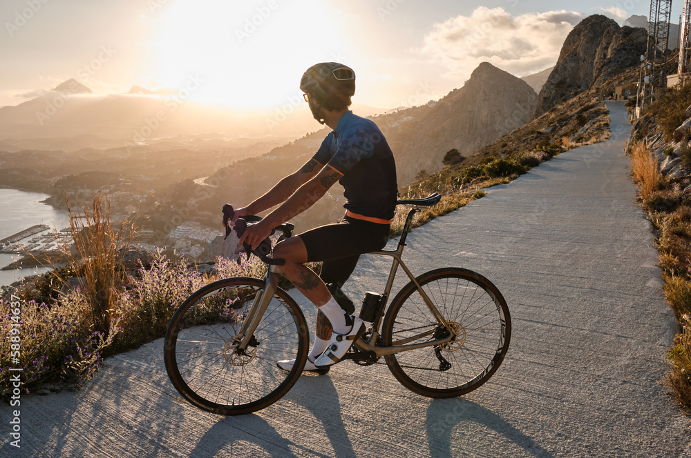 Fototapeta premium A male cyclist enjoying a beautiful seaside view during sunset amidst her training.Man cyclist wearing blue and black cycling kit and black helmet on a gravel bike.Calp,Alicante,Spain