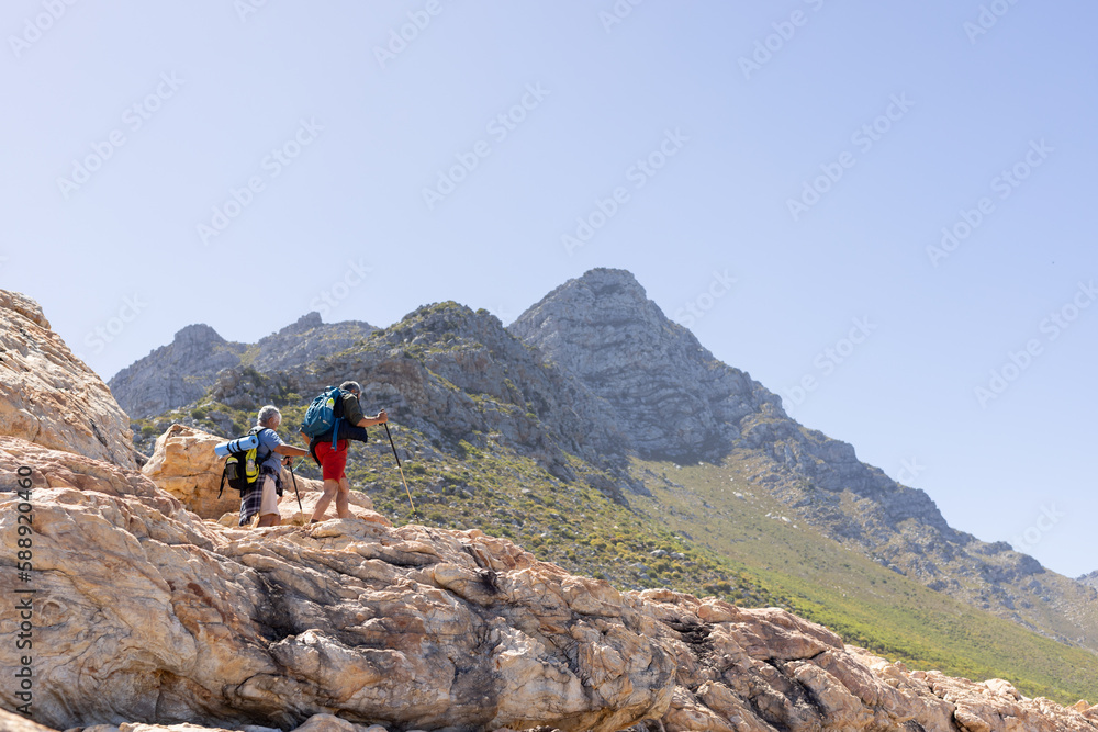 Happy senior biracial couple wearing backpacks, hiking with trekking poles, with copy space