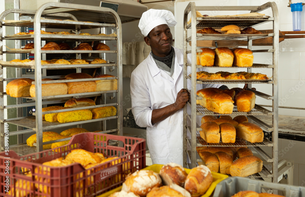 Professional African American baker carrying fresh baked bread on tray rack trolley in bakery workshop ..