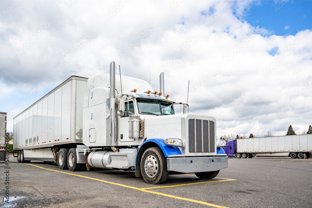Stylish white big rig semi truck with blue accents and loaded dry van semi trailer standing on the market spot on truck stop parking lot for truck driver rest