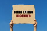 Binge eating disorder text on box paper held by 2 hands with isolated blue sky background. This message board can be used as business concept about compulsive overeating.