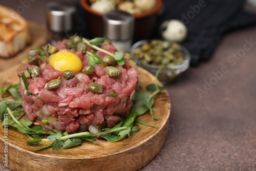 Tasty beef steak tartare served with yolk, capers and microgreens on brown table, closeup. Space for text
