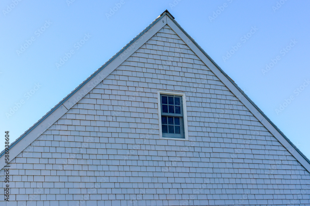 The exterior peak of a white wooden house with black shingles, a vent pipe and two windows. The sky in the background is blue with some clouds. There trees on both sides of the residential structure