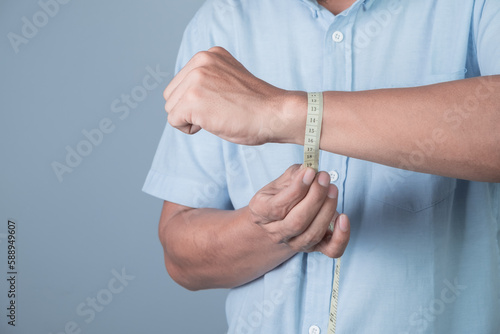 A man measures his wrist with a tape measure on light blue background, weight loss concept