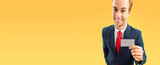 Funny businessman in grey suit and red tie, showing blank business or plastic credit card, isolated on yellow-orange background. Wide banner image.