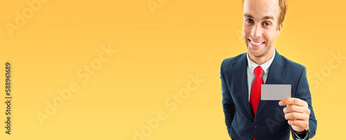 Funny businessman in grey suit and red tie, showing blank business or plastic credit card, isolated on yellow-orange background. Wide banner image. photo