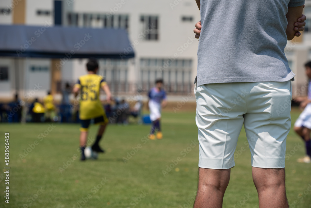 Father standing and watching his son playing football in a school tournament on a clear sky and sunny day. Sport, outdoor active, lifestyle, happy family and soccer mom and soccer dad concept.