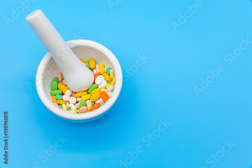 A mortar is placed on a blue background and filled with colorful pills capsules, a pestle, and an empty space to use as a copyspace.