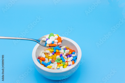 A mortar is placed on a blue background and filled with colorful pills capsules, a spoon and an empty space to use as a copyspace. Someone is floating with a spoon