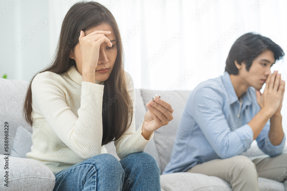 Stressed asian young couple man, woman quarrel on couch, relationship in trouble. Wife's hand holding wedding ring in disappointment and upset her husband, which may lead to divorce. Problem of family Stressed asian young couple man, woman quarrel on couch, relationship in trouble. Wife's hand holding wedding ring in disappointment and upset her husband, which may lead to divorce. Problem of family