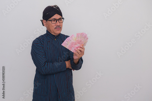 A Javanese man wearing traditional outfit holds some money; serious expression. photo