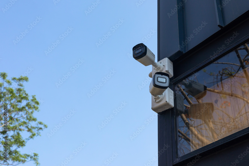 Two white surveillance cameras on black pillars in the garden near buildings on blue background. outdoor security cctv camera Two white surveillance cameras on black pillars in the garden near buildings on blue background. outdoor security cctv camera