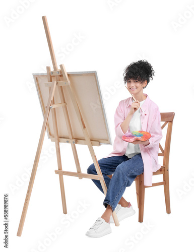 Young woman holding brush and artist`s palette near easel with canvas against white background
