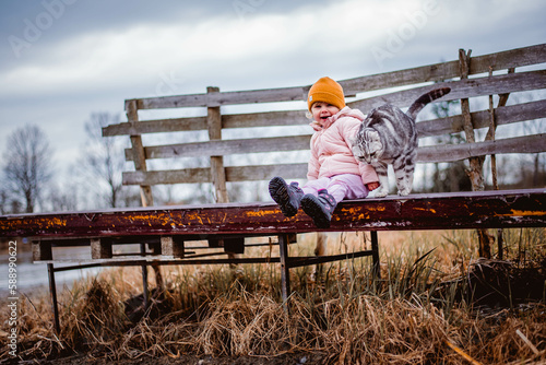two year old laughing girl in yellow hat sitting on pier by empty pond, british whiskas cat rubs and purrs on her, happy child with beloved pet, kids and pets, outdoor kid 