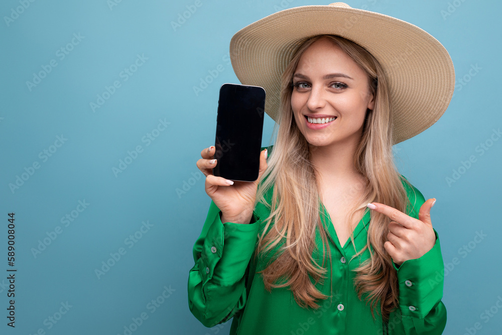 horizontal photo of charming woman on vacation in summer straw hat showing phone screen with mocup on blue studio background horizontal photo of charming woman on vacation in summer straw hat showing phone screen with mocup on blue studio background