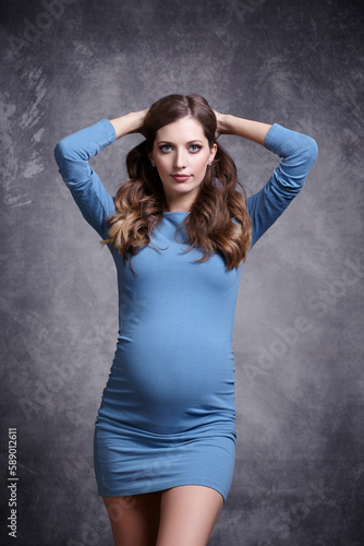 Portrait of young pretty pregnant woman on gray studio background. Female in blue dress with hands behind head.