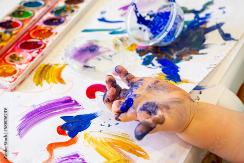 Toddler paints hand with colorful watercolor paint while painting on white paper