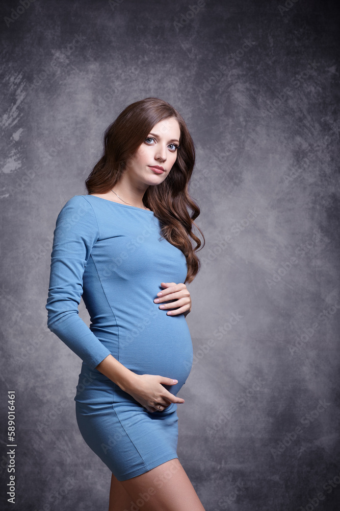Portrait of young pretty pregnant woman on gray studio background. Female in blue dress with hands near pregnant belly Portrait of young pretty pregnant woman on gray studio background. Female in blue dress with hands near pregnant belly