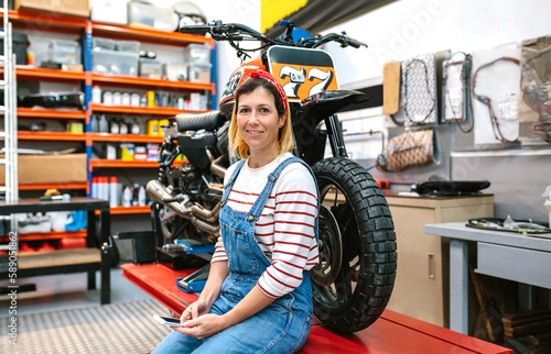 Portrait of smiling mechanic woman looking at camera while holding phone in her hands sitting over platform with custom motorcycle on factory