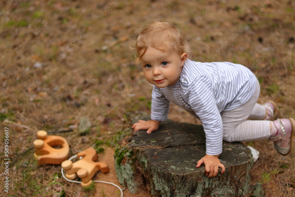 Outdoor shot of cute funny infant baby girl with blonde hair wearing striped shirt playing with wooden toys, looking away, climbed a stump in the forest. Outdoor shot of cute funny infant baby girl with blonde hair wearing striped shirt playing with wooden toys, looking away, climbed a stump in the forest.