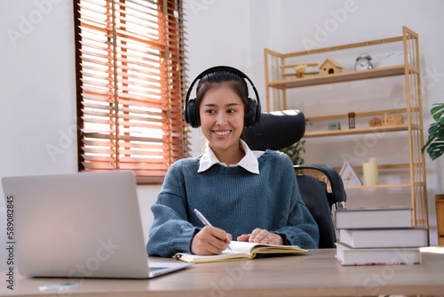 Candid young attractive asian female employee wear earphone listen to digital online course development program in quarantine social distance at home reskill upskill school class for career path.