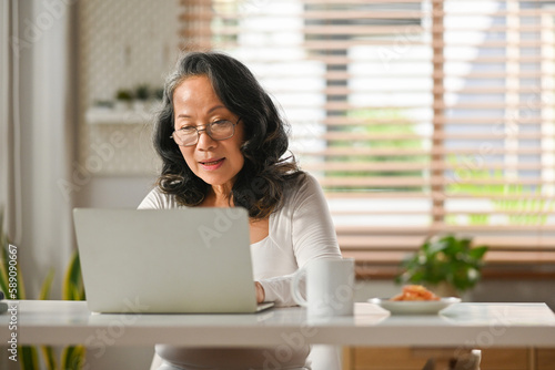 Beautiful senior woman entrepreneur working remote from home, reading email on laptop computer