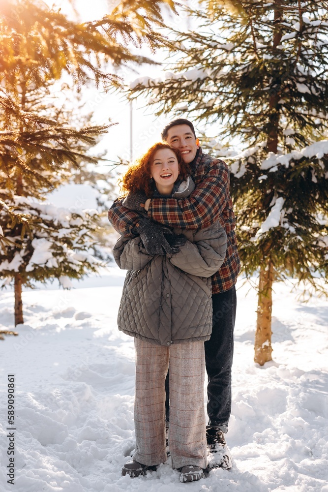 A young man hugs his red-haired girlfriend by the shoulders. Young people in love pose standing against the backdrop of a snow-covered forest. A married couple walks in nature on a winter sunny day. A young man hugs his red-haired girlfriend by the shoulders. Young people in love pose standing against the backdrop of a snow-covered forest. A married couple walks in nature on a winter sunny day.