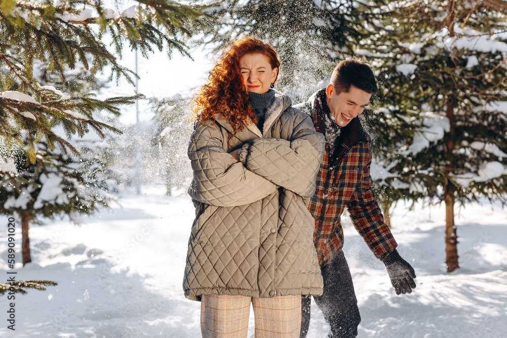 A young brunette sprinkles snow on his red-haired girlfriend. A man teases his girlfriend while walking in a winter sunny forest.
