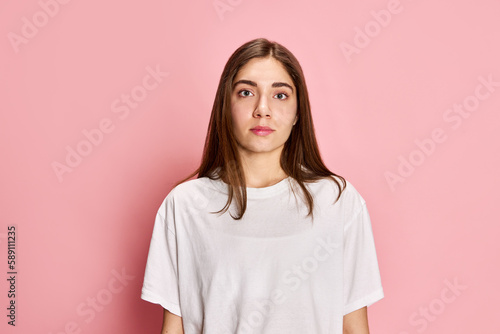 Portrait of young brunette girl in white-t-shirt attentively looking at camera against pink studio background. Casual style. Concept of youth, emotions, facial expression, lifestyle