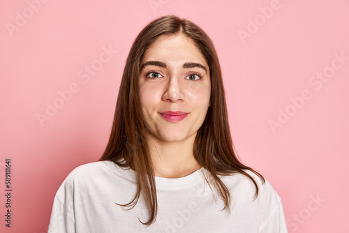 Portrait of beautiful young girl in white t-shirt looking at camera with little smile  posing against pink studio background. Concept of youth  emotions  facial expression  lifestyle
