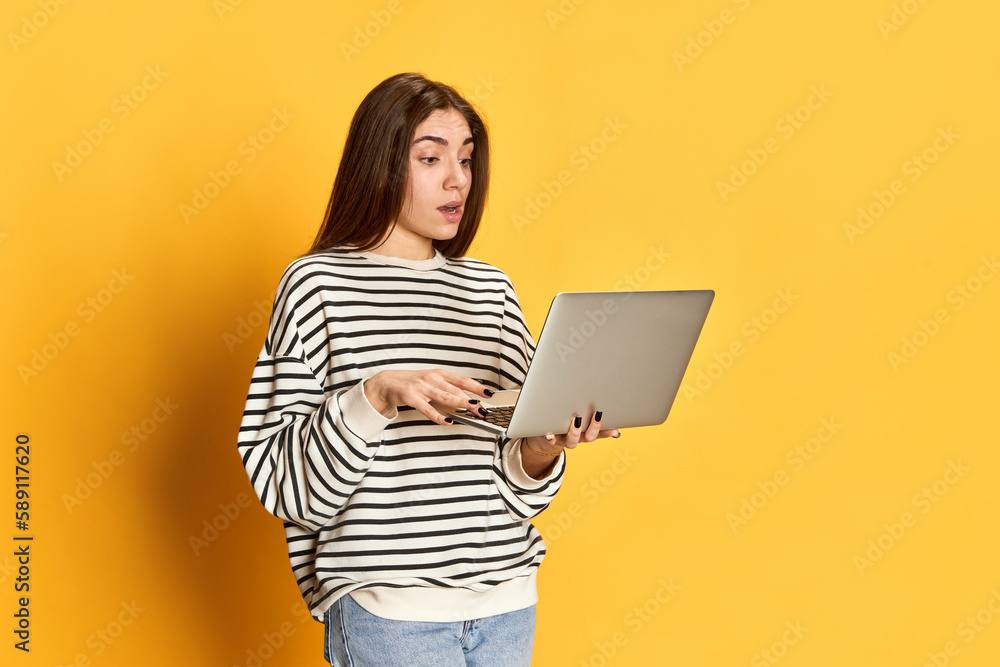 Portrait of young girl in striped sweater using laptop for working and studying, posing againt yellow studio background. Freelance job. Concept of youth, emotions, facial expression, lifestyle Portrait of young girl in striped sweater using laptop for working and studying, posing againt yellow studio background. Freelance job. Concept of youth, emotions, facial expression, lifestyle